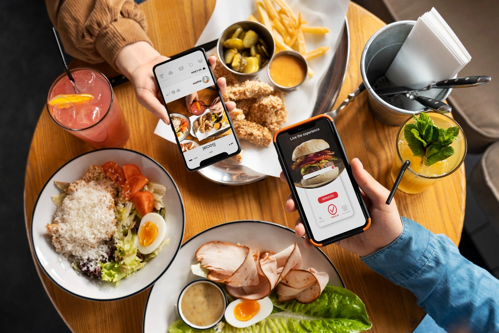 Two people at a table with salad, sliced turkey, fries, pickles, and drinks browse food delivery apps on their smartphones, showing images of burgers and pizzas.